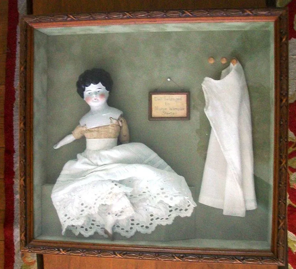 Antique porcelain doll in a white dress displayed in a shadow box beside her gown